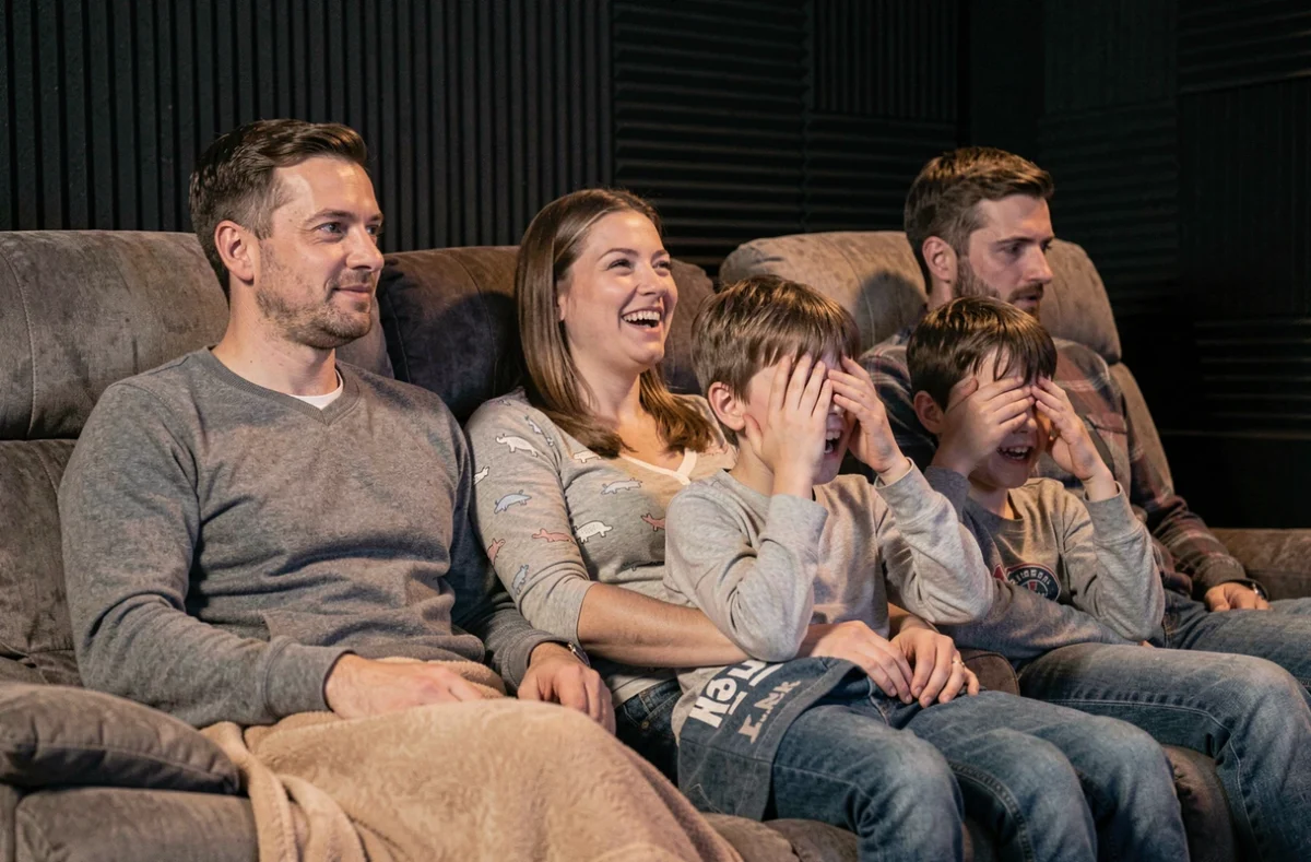 Famille de quatre personnes réagissant différemment devant un film dans une salle de cinéma maison, entre rires et frayeur amusante